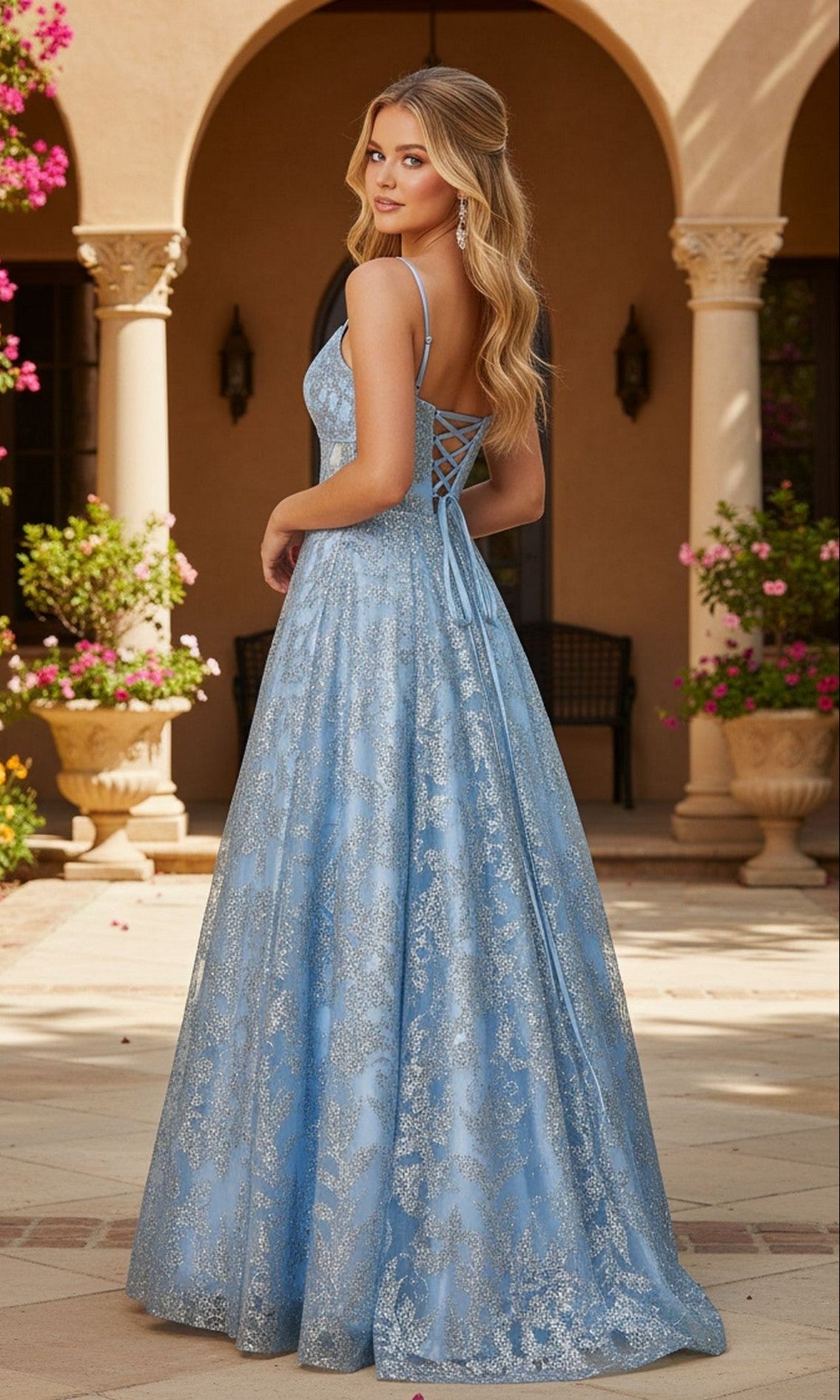 A woman in the Blondie Nites 5985BN long prom dress, featuring a light blue hue, lace-up back, and glitter details, stands outdoors facing away by stone columns and flowering plants.